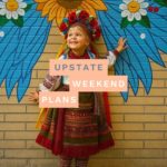 Little girl dressed in traditional Ukrainian clothes with a mural backdrop and with the "Upstate Weekend Plans" text written across the Hudson Valley, NY photo.