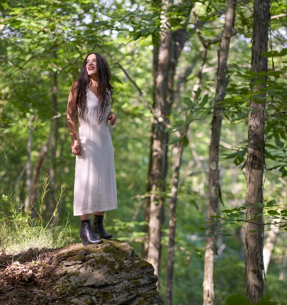 Rachael Yamagata in the woods standing on a large rock in Woodstock NY