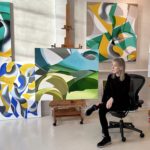 In the Studio With Ginnie Gardiner sitting on a chair in her studio with her paintings all around