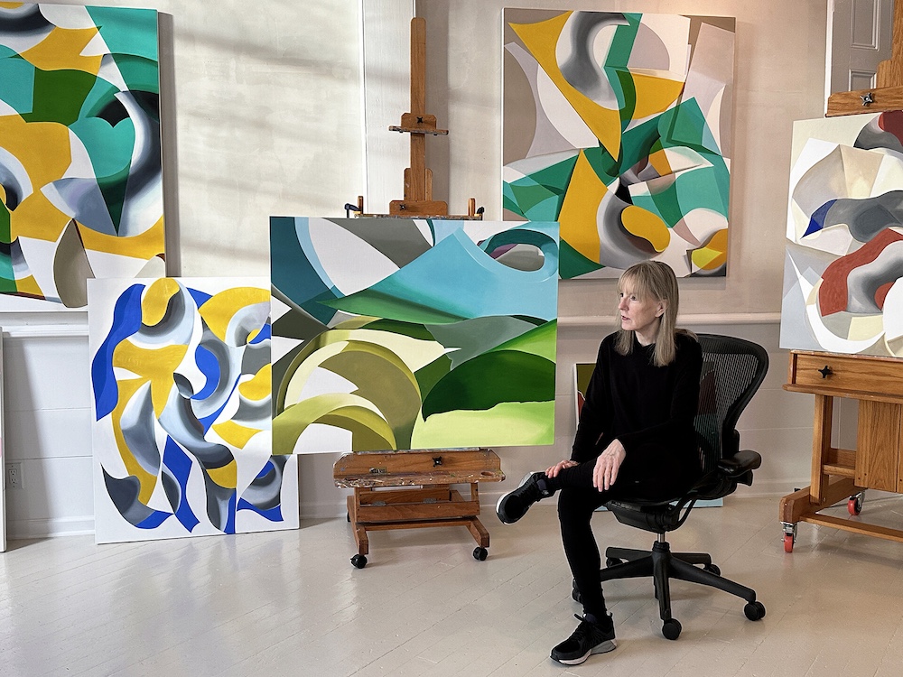 In the Studio With Ginnie Gardiner sitting on a chair in her studio with her paintings all around