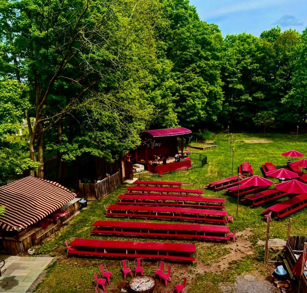 Aerial view of the Garden at Colony Woodstock showing red picnic tables, umbrellas and a stage with grass and trees all around.