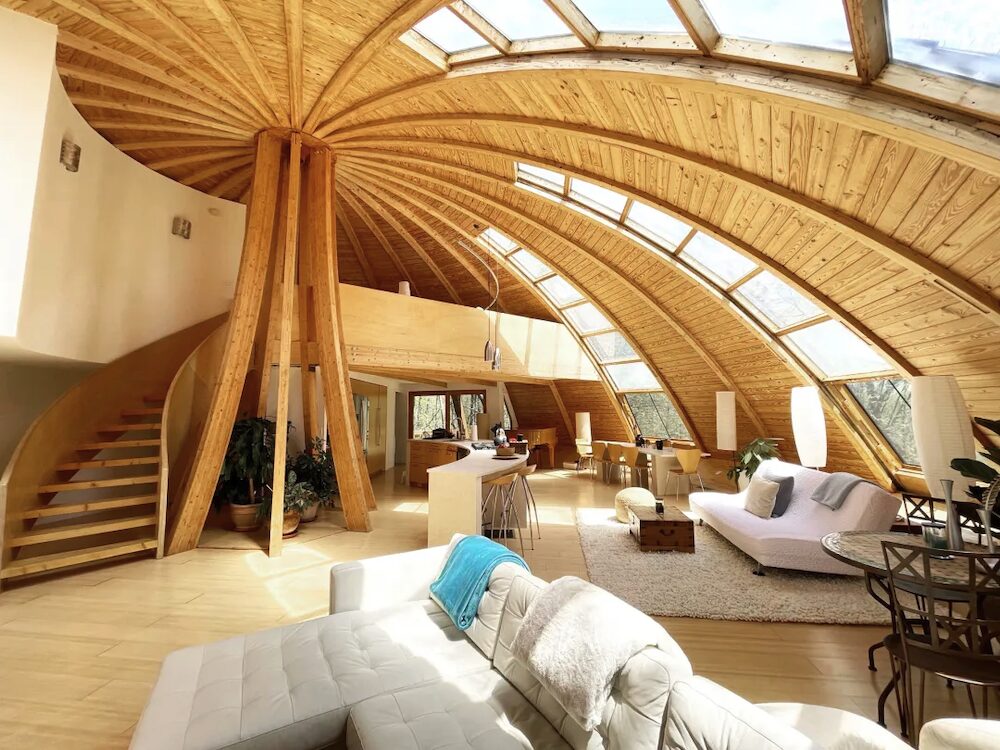 Living room filled with couches, staircase, with a dome shaped ceiling and strips of windows between panels.