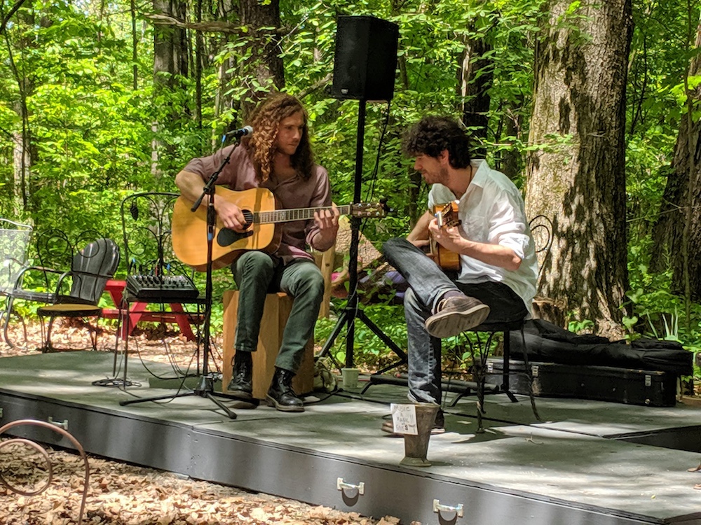 two men playing guitars on a makeshift stage in the woods at a the Rail Trail Cafe