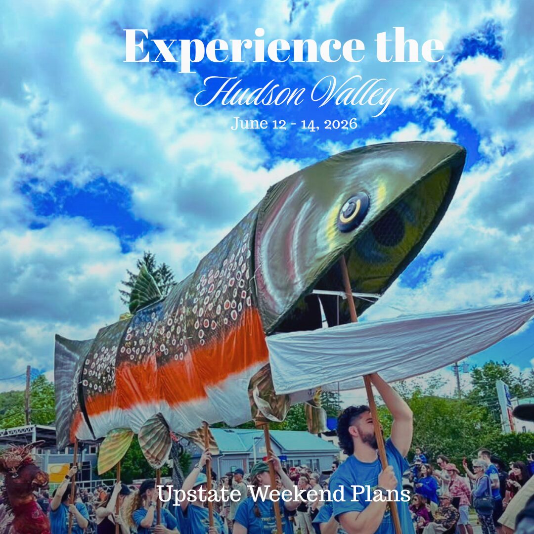 A group of people with blue shirts holding an oversized trout puppet in the air as they walk through the trout parade in Livingston Manor with the "Experience the Hudson Valley" and "Upstate Weekend Plans" text written across the photo for this upcoming weekend dates.