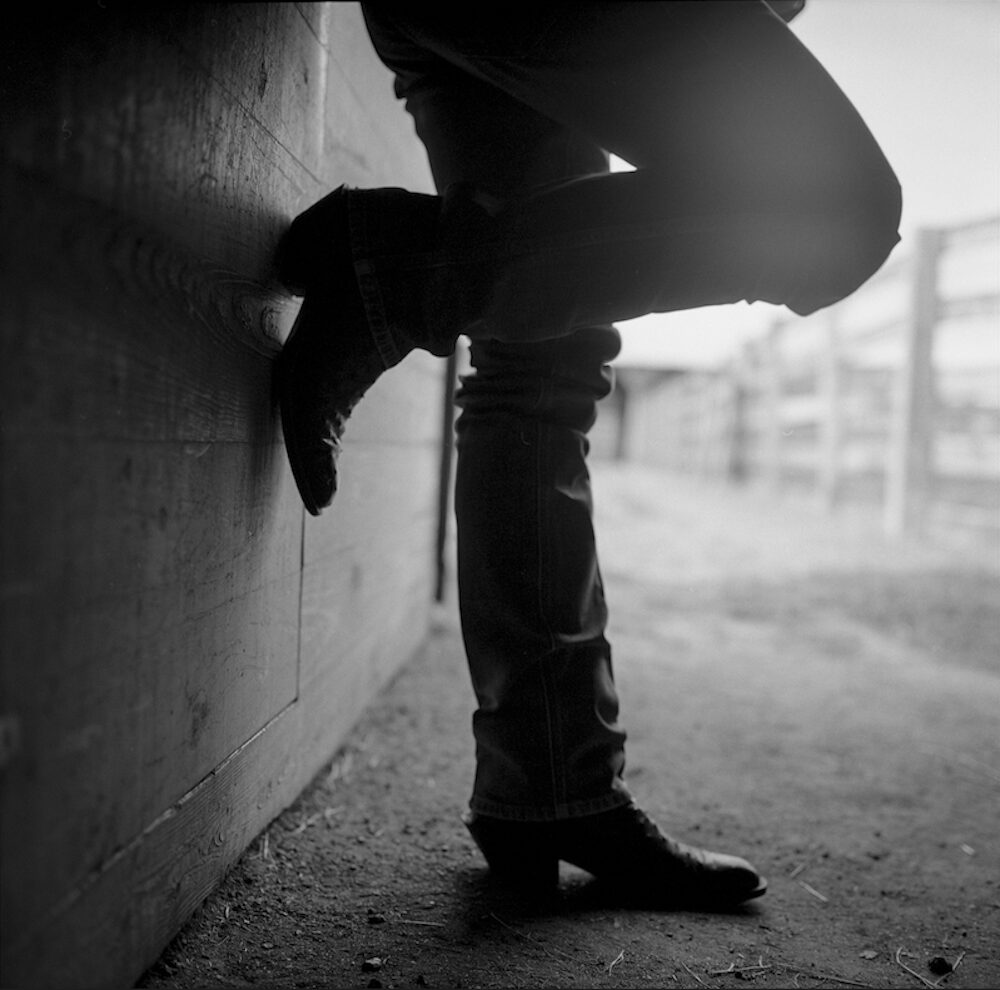 Lyle Lovett leaning against a wall with one leg up against the wall. POV is from the side and only his legs.
