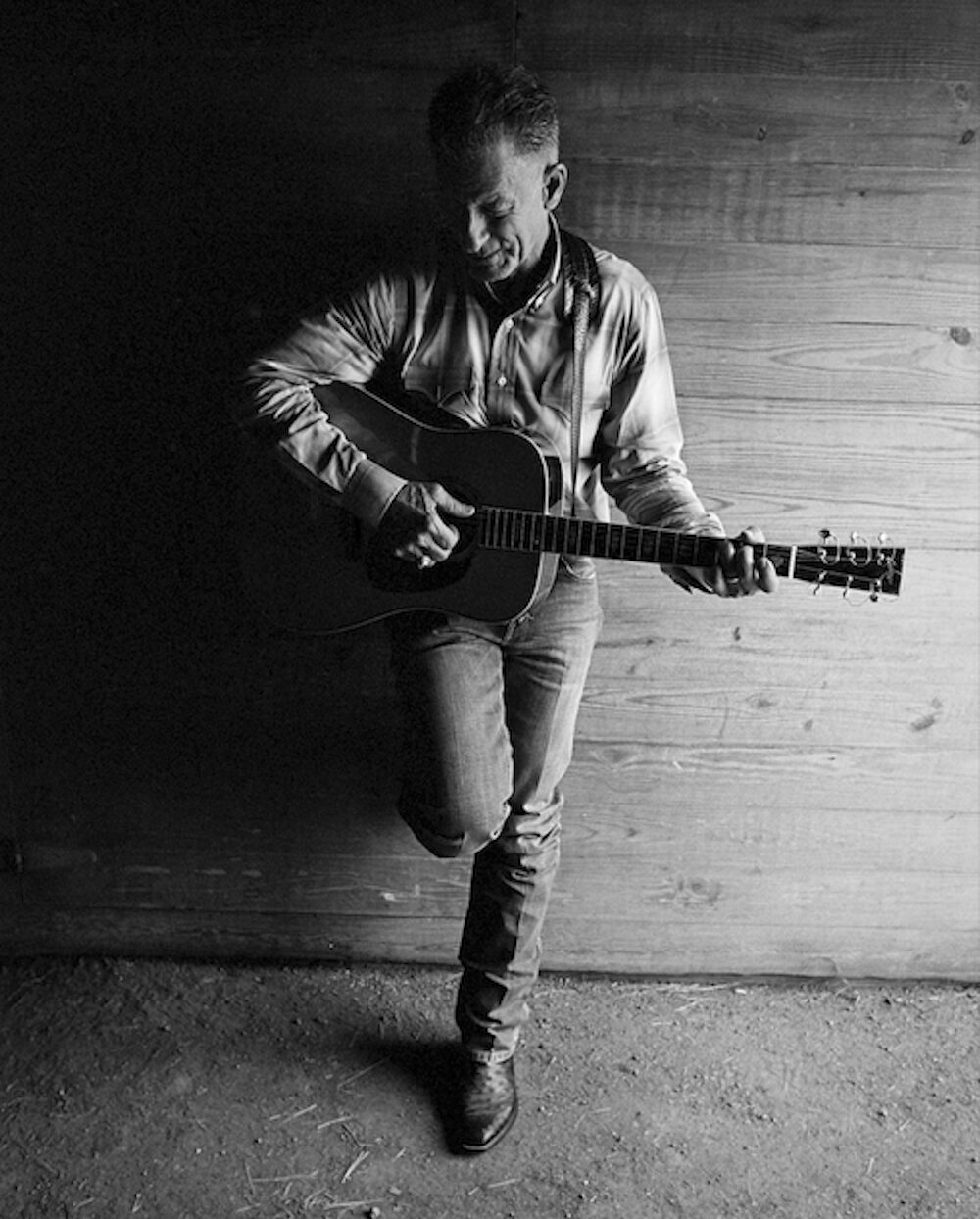 Lyle Lovett leaning against a wall with one leg up against the wall with his guitar.