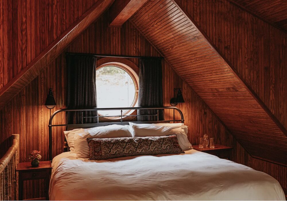 A bed against the wooden walls with a circle window at Deer Mountain Inn in Tannersville NY.