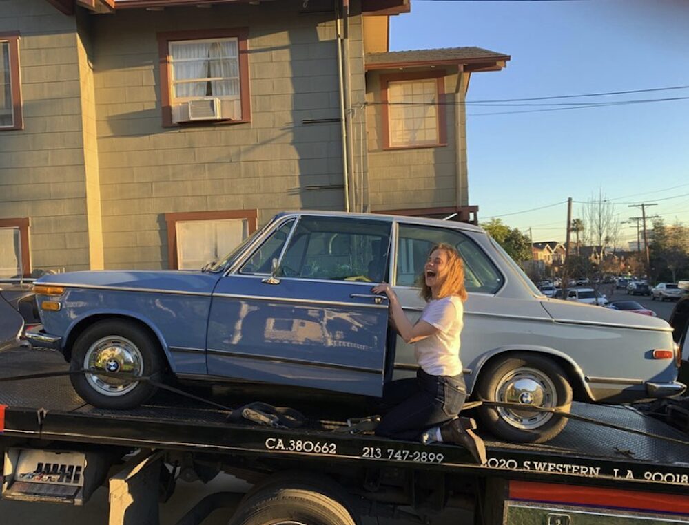 Fiona Dourif laughing halfway in the door of a blue BMW on a car bed.