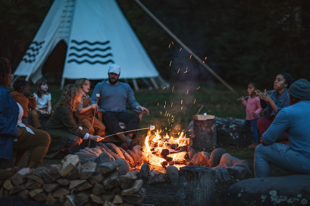 Families gathered around a firepit roasting marshmellows with a tipi in the background at Graham & Co in Phoenicia NY.