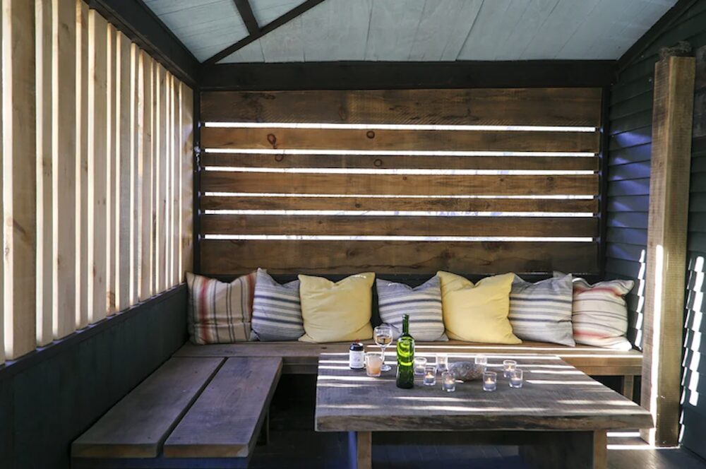 Wooden cabin benches with pillows and coffee table with bottle of wine at The Pines in Mount Tremper NY.