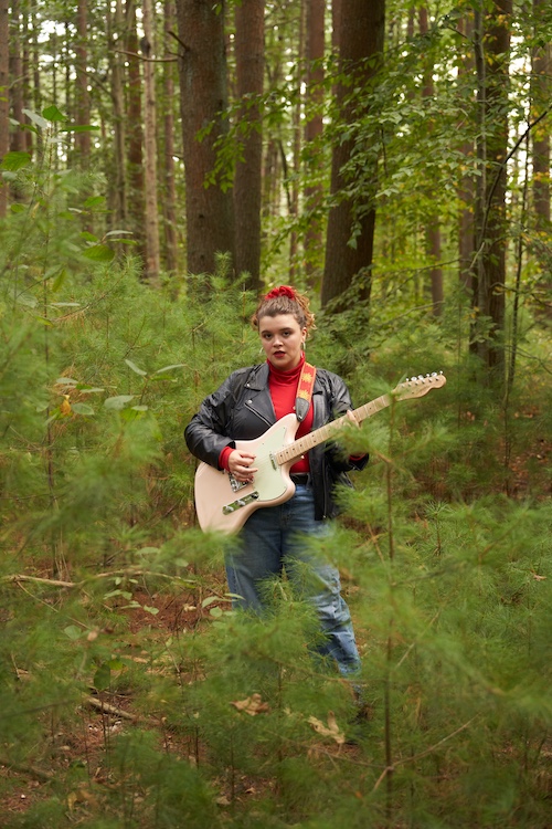 Xondra holding her guitar in the woods with trees all around her.